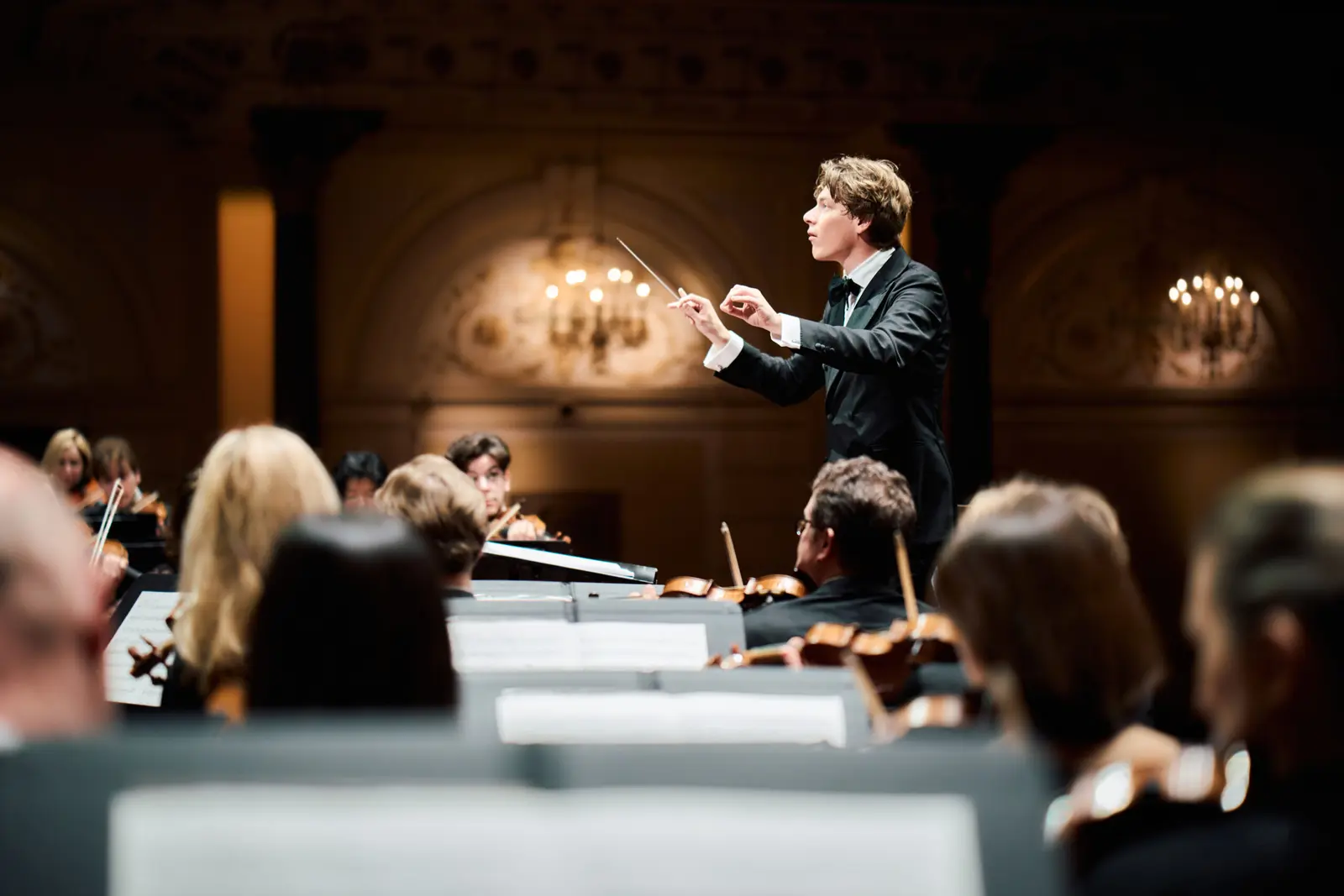 A conductor leads an orchestra in a concert hall.