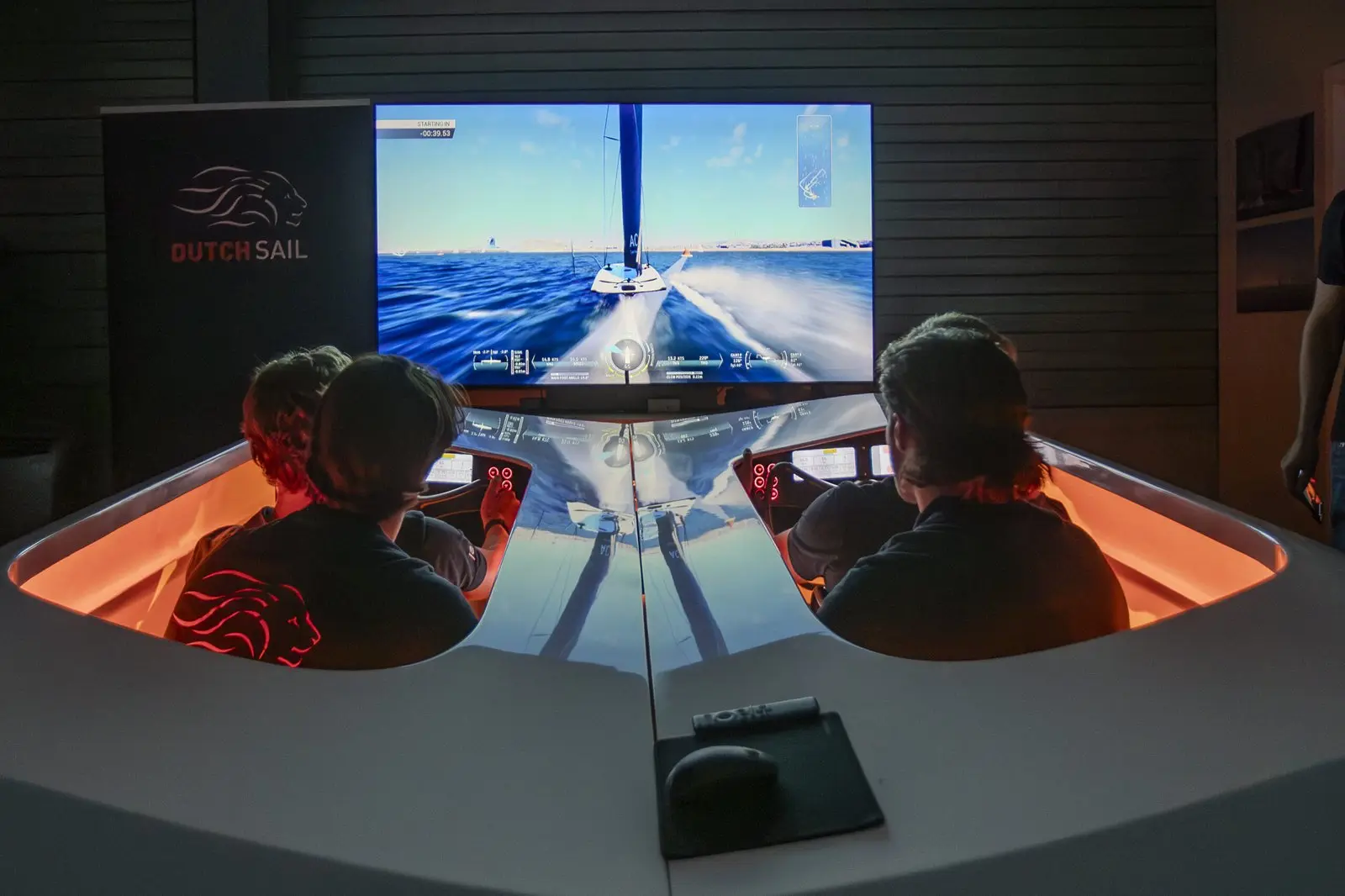 Twee mensen in een zeilsimulator kijken naar een scherm met een zeilboot.