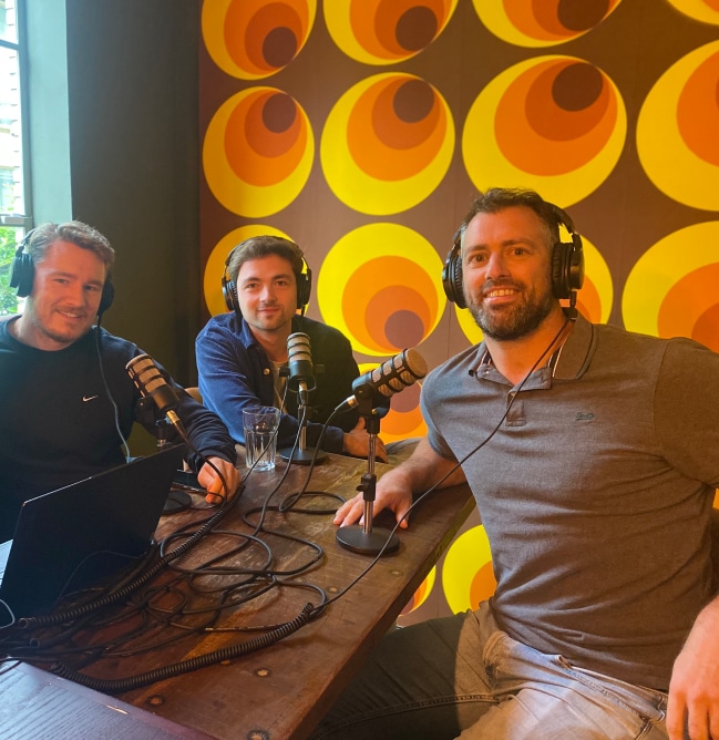 Três criadores do podcast De Databar estão sentados à mesa, com auscultadores, atrás de microfones.